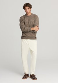 Maglione lavorato a maglia marrone con scollo rotondo, abbinato a pantaloni tapered color beige e scarpe marroni con lacci, su uno sfondo neutro.