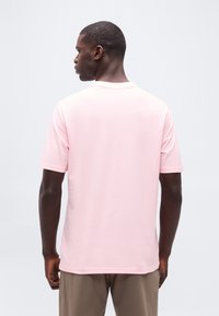 Άνδρας που κοιτάζει προς τα πίσω, φορώντας ανοιχτό ροζ μπλουζάκι με κοντά μανίκια και παντελόνια ταουπ, με φόντο ένα απλό ανοιχτόχρωμο τοπίο.