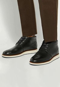 Black leather chukka boots with white soles, featuring a smooth finish, lace-up front, and contrasting brown accents on the sole edge.