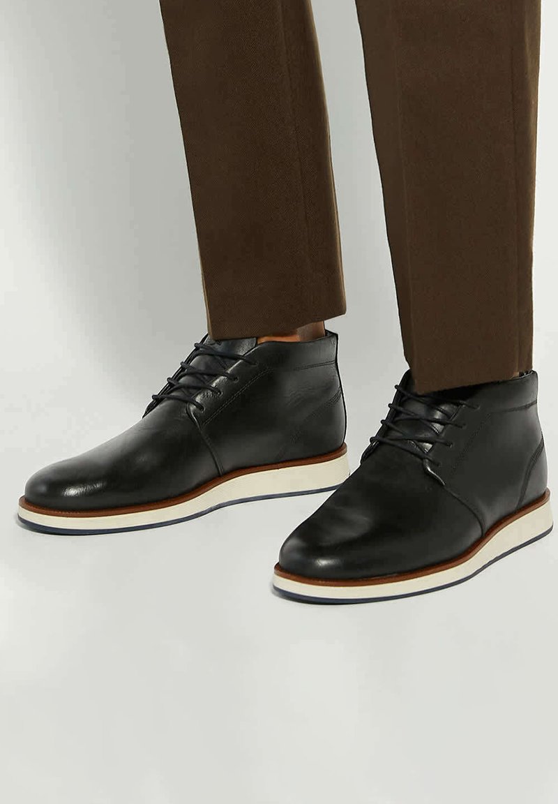 Black leather chukka boots with white soles, featuring a smooth finish, lace-up front, and contrasting brown accents on the sole edge.