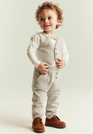 Peuter met krullend haar, gekleed in een beige vest, bijpassende broek, wit shirt met lange mouwen en bruine schoenen, staand tegen een lichte achtergrond.