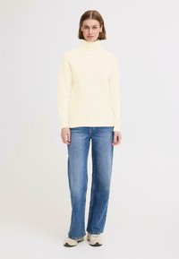Ljusgul ribbad polotröja, åtsittande med lång ärm, ihop med blå vida jeans och beige sneakers. Enfärgad bakgrund.