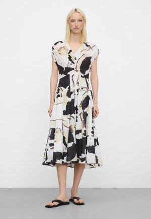 Vrouw draagt een midi-jurk met abstracte print in zwart, wit en beige, met vleermuismouwen en zwarte platte sandalen, staande tegen een witte achtergrond.