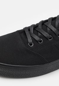 Sneaker noir en toile avec un dessus texturé, lacets noirs plats et œillets ronds. La semelle en caoutchouc présente un motif antidérapant pour une meilleure adhérence.