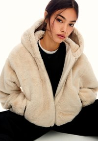 Veste en fausse fourrure couleur crème avec fermeture éclair sur le devant, capuche et poches latérales. Texture douce et forme carrée, superposée sur une chemise noire.