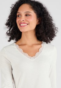 Lichtbeige langetermijn shirt met V-hals en kantafwerking rond de halslijn, gemaakt van zachte stof met een gladde textuur.