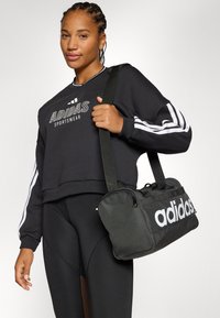 Osoba s pletenicama nosi crnu Adidas duksericu i tajice, držeći crnu sportsku torbu s vidljivo prikazanim Adidas logom.