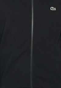 Veste noire zippée avec une fermeture éclair argentée visible et un petit logo de crocodile vert sur la partie supérieure gauche de la poitrine.