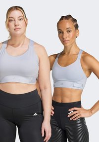 Light gray sports bras with adjustable straps, paired with black leggings featuring a subtle wave pattern and logo. Smooth fabric texture.