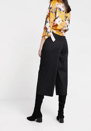 Vrouw die een gele blouse met bloemenprint draagt, zwarte wijde cropped broek en zwarte laarzen met hakken, staand tegen een effen witte achtergrond.