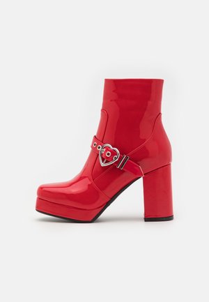 Bottine à plateforme rouge brillante avec talon épais et sangle à boucle argentée en forme de cœur sur fond blanc.