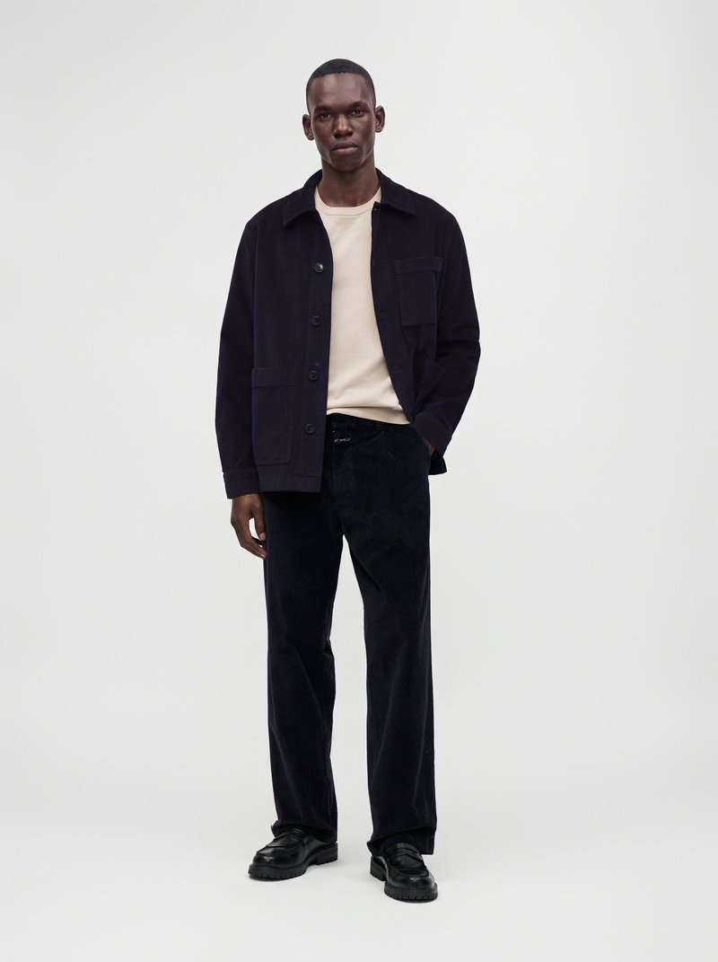 Homme debout portant une chemise beige, une veste noire boutonnée, un pantalon noir et des chaussures noires sur un fond clair uni.
