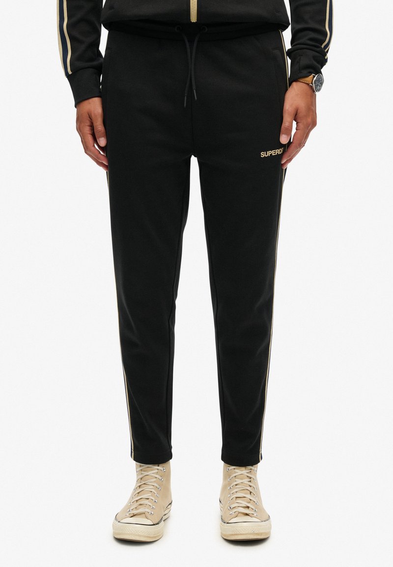 Man in zwarte getailleerde sweatpants met gouden zijkanten en het 'Superdry' logo, gecombineerd met beige hoge sneakers en een zwart horloge.