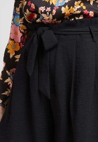 Jupe noire texturée avec une ceinture nouée à la taille. Le tissu semble léger et fluide, complétant un haut avec un motif floral.