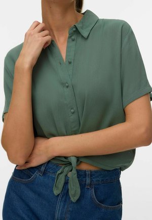 Chemise verte à manches courtes boutonnée, nouée à la taille, portée avec un jean bleu taille haute, la main du modèle touche le col et le bras est croisé sur le torse.