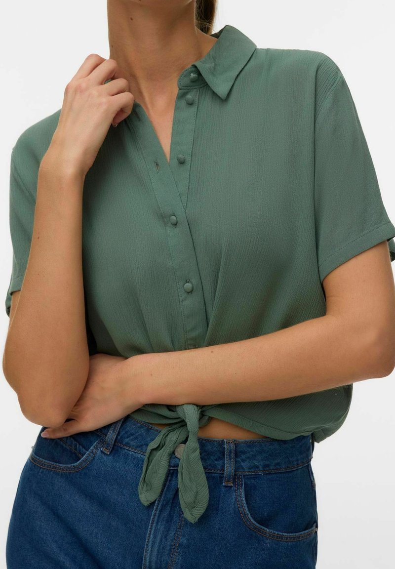 Chemise verte à manches courtes boutonnée, nouée à la taille, portée avec un jean bleu taille haute, la main du modèle touche le col et le bras est croisé sur le torse.