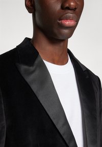 Sacou din catifea neagră cu reveruri din satin, purtat peste un tricou alb. Prim-planul arată detaliile gulerului și textura materialului.