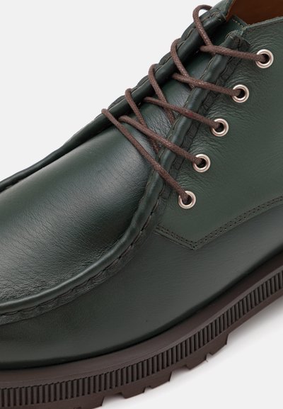 Green leather shoe with brown laces, featuring silver eyelets, a textured toe cap, and a corrugated black sole.