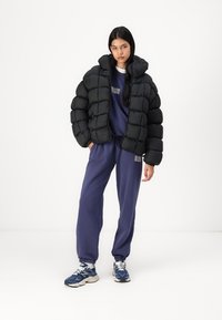 Svart pufferjakke med hette, med en quiltet design. Kombinert med en lilla genser og matchende joggebukser. Hvite sneakers.
