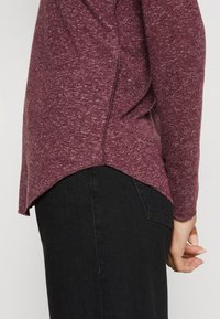 Maglietta bordeaux a maniche lunghe con una texture mélange, orlo asimmetrico, dettaglio sulla cucitura laterale, indossata sopra jeans neri, con posizionamento delle mani visibile.