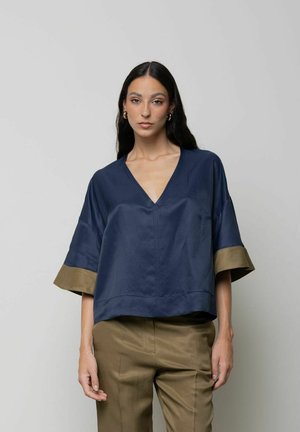 Donna con capelli lunghi e scuri indossa una maglia blu navy con scollo a V, maniche larghe e polsini tan, abbinata a pantaloni tan coordinati su sfondo uniforme.
