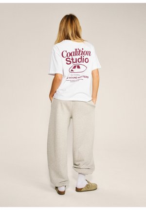 Kobieta z długimi blond włosami, ubrana w białe skarpetki, beżowe buty wsuwane, szare spodnie dresowe i białą koszulkę z czerwonym tekstem i grafiką "Coalition Studio".