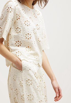 Femme portant un haut en tissu ajouré blanc et une jupe assortie avec des motifs floraux découpés, une main dans la poche, debout devant un fond uni.