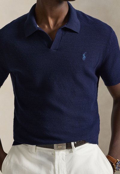 Σκούρο μπλε polo με κοντά μανίκια, υφή ύφανσης και γιακά. Διαθέτει κεντημένο λογότυπο στο αριστερό στήθος. Φοριέται με λευκό παντελόνι.