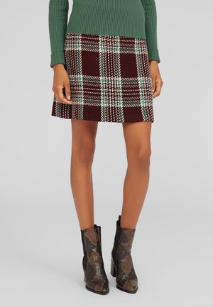 Femme portant un pull vert, une jupe trapèze à carreaux bordeaux et blancs, et des bottines marron à motif serpent, debout.