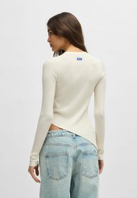 Top a maniche lunghe a costine di colore crema con scollatura rotonda e orlo asimmetrico. Abbinato a jeans in denim blu chiaro, con tasche posteriori.