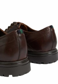 Zapatos de cuero marrón con un acabado texturizado, que cuentan con un tacón bajo, suela de goma oscura y una tab de talón a rayas en verde y azul.