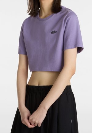 Kobieta ubrana w fioletowy, przycięty t-shirt Vans i czarne spodnie z elastycznym pasem, stoi z rękami swobodnie złączonymi.