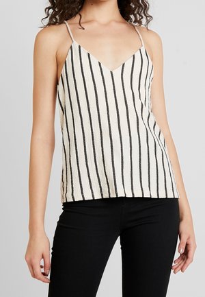 Vrouw draagt een wit mouwloos topje met zwarte verticale strepen en een zwarte broek, staand tegen een effen lichte achtergrond.