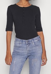 Kvinna klädd i en svart, knappad topp med halvärmar, instoppad i ljusblå, högmidjade jeans med slitningar och framfickor.