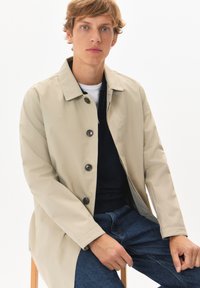 Bežna vodoodporna jakna s ovratnikom in temnimi gumbi, oblečena čez mornarsko modro pulover, v kombinaciji z modrimi kavbojkami.