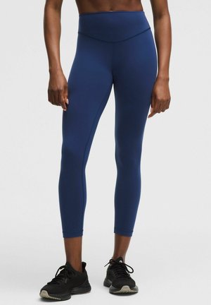 Person iført højttaljede marineblå leggings og sorte træningssko, stående mod en ensartet lysegrå baggrund.