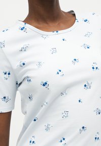 Gros plan d'une personne portant une chemise blanche à manches courtes avec un motif floral bleu et un col rond.