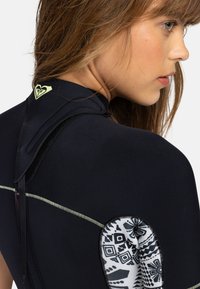 Une combinaison de plongée noire avec un panneau en néoprène décoratif à motif sur l'épaule gauche. Présente un logo néon sur le col et des coutures contrastées.