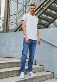 Camiseta azul claro de estilo tie-dye, jeans de mezclilla y zapatillas Converse grises, de pie en unos escalones de piedra con una barandilla de metal texturizada y un fondo.