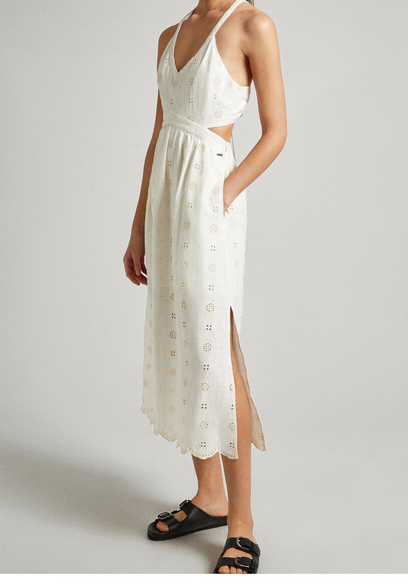 Femme portant une robe midi blanche sans manches avec un motif en broderie anglaise, des découpes sur les côtés, une fente sur un côté, et des sandales noires à boucle.