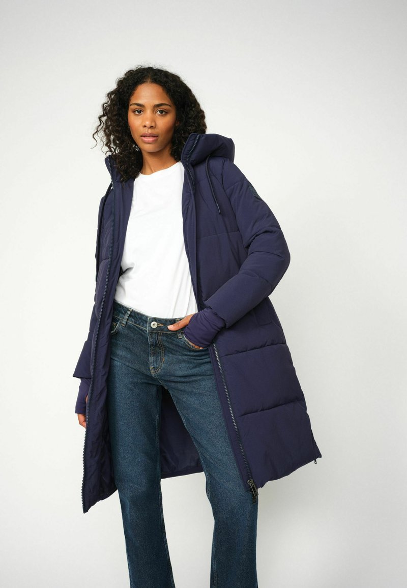 Mornarska puhasta jakna s kapuco in vertikalnim prešitim vzorcem, v kompletu z belim T-shirtom in modrimi traper hlačami. Ima zadrgo in nastavljive vrvice.