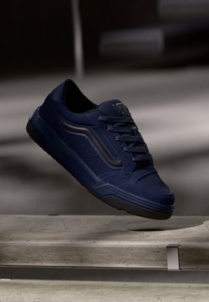 Baskets bleu marine avec des accents noirs et des lacets, présentées en suspension au-dessus d’un banc en béton dans un environnement intérieur faiblement éclairé.