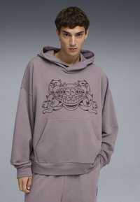 Sudadera púrpura con un diseño gráfico de gatos y un motivo de dientes. Hecha de una tela suave con un corte relajado y un bolsillo frontal.