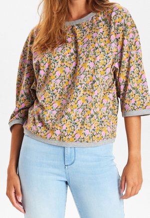Femme portant un pull ample à motif floral avec des couleurs jaune, rose et verte, associé à un jean bleu clair sur fond blanc.