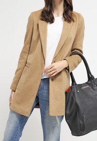 Kabát z vlněné směsi v barvě velbloudí srsti s klopami s výřezy a dvěma kapsami, doplněný o roztrhané modré džíny a černou koženou kabelku s červenými detaily.