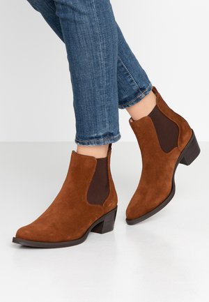 Brown suede ankle boots with elastic side panels worn with blue denim jeans on white floor.