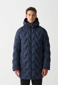 Muž v dlhom námorníckom prešívanom puffer kabáte s kapucňou, predným zipsom a zipsovými bočnými vreckami, stojaci na jednolatom pozadí.