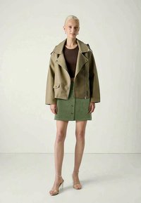 Veste en simili cuir vert olive avec zip asymétrique, associée à un haut marron et une jupe en velours côtelé verte avec des boutons sur le devant. Talons transparents.