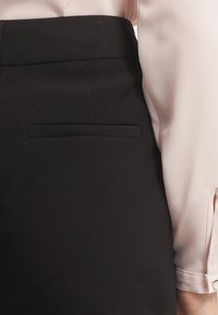 Detailní záběr na černou přizpůsobenou sukni s kapsou na zadní straně, doplněnou o světle růžovou halenku s dlouhým rukávem, která je na zápěstí ohrnutá.