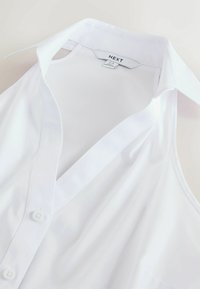 Chemise blanche sans manches avec col, boutons et une étiquette de vêtement indiquant "NEXT" cousue à l’intérieur de l’encolure.